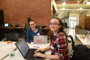 Zwei Frauen sitzen an einem Tisch mit Laptops, einer Flasche, einem Stift und Papier in einem Büro mit einer roten Backsteinwand, Deckenlampen, einer Treppe und einer Eingangstür im Hintergrund.