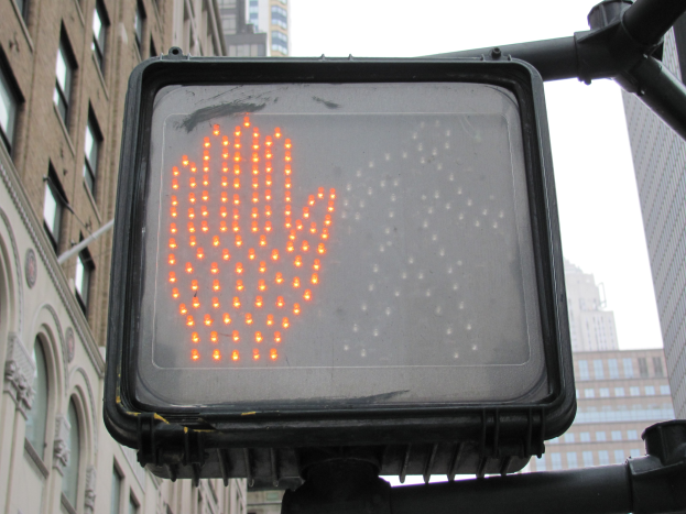 Eine Ampel mit rotem Handsymbol in einer städtischen Umgebung mit Gebäuden auf beiden Seiten und einem klaren Himmel darüber.