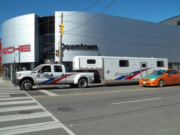 Polizeifahrzeug vor einem Gebäude mit sichtbarem Text geparkt, Verkehrssignal mit Stange und Dröhnen im Vordergrund, Bäume und klarer blauer Himmel im Hintergrund.