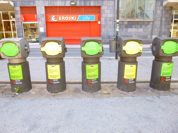 Eine Reihe von Parkautomaten entlang einer Straße mit einem Gebäude, das Glasfenster und eine Rolllade mit Text aufweist, begleitet von einem nahen Müllcontainer.