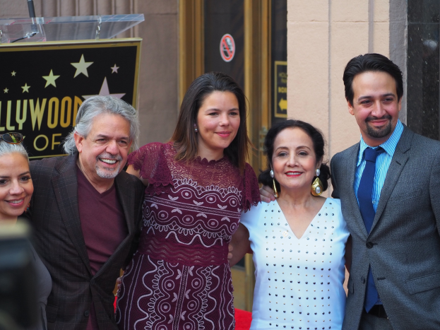 Gruppe von Menschen, die auf dem roten Teppich der Zeremonie des Hollywood Walk of Fame lächeln und posen, mit einer Tafel, einem Glaspodium und einer Wand im Hintergrund.