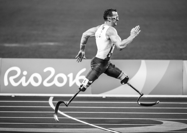 Schwarzes und weißes Foto eines Mannes mit einer Prothese, der auf einer Laufbahn läuft, mit einer Tafel im Hintergrund, auf der "Rio 2016 Paralympic Games" steht.
