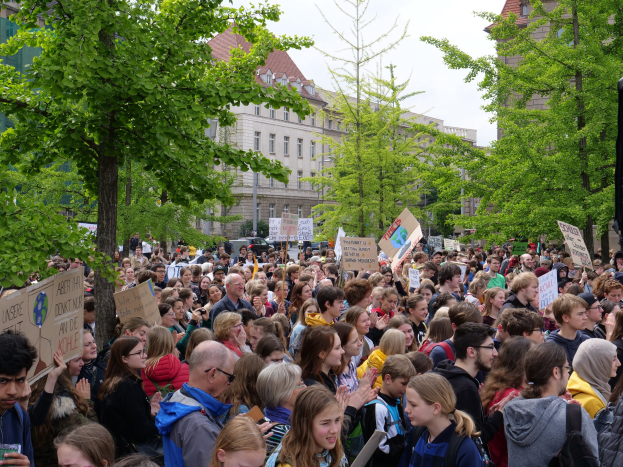 Große Menge von Menschen protestiert vor einem Gebäude in Berlin, mit Schildern, Bäumen, Fahrzeugen, einem Lautsprecher und Himmel.