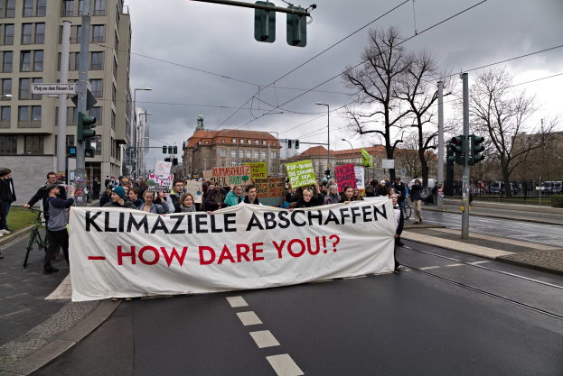 Eine Gruppe von Menschen marschiert mit einem Banner, auf dem "Klimaziele Abschaffen - Wie wagst du es?" steht, die Straße entlang, mit einem Radfahrer im Vordergrund und Gebäuden, Bäumen und Verkehrszeichen im Hintergrund unter einem klaren blauen Himmel.