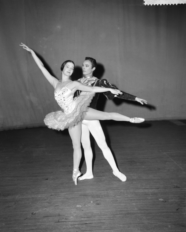 Schwarzes und weißes Foto eines Mannes in einem weißen Hemd und schwarzen Hosen und einer Frau, die auf der Bühne tanzt, mit einem Vorhang als Hintergrund, ihre Gliedmaßen sind in perfekter Harmonie verschlungen.