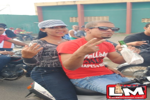 Ein Mann sitzt auf einem Fahrrad und hält eine Bierflasche in der Hand, mit einem Mädchen hinter ihm und vielen anderen Menschen auf Fahrrädern im Hintergrund, vor einer Wand.