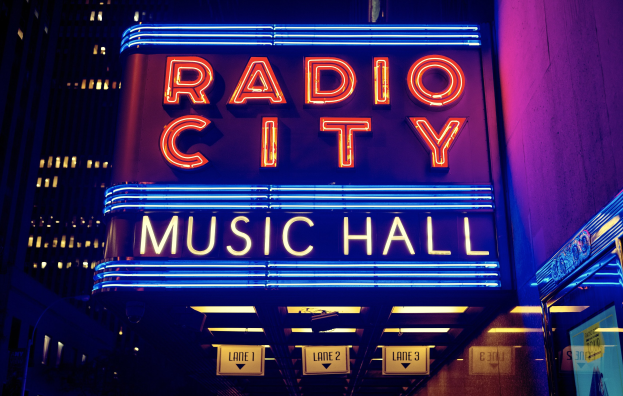 Radio City Music Hall in New York City, hell erleuchtet in der Nacht, umgeben von hohen Gebäuden und einem "Radio City"-Schild auf der rechten Seite.