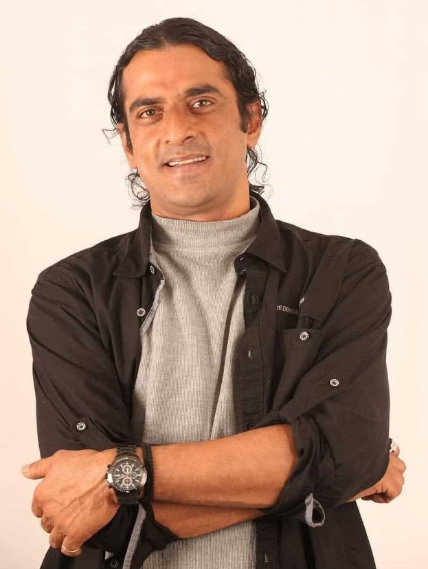 Ein Mann in einem schwarzen und aschfarbenen Kleid mit verschr├Ąnkten Armen, der eine Uhr tr├Ągt und gegen einen weißen Hintergrund steht, in Gedanken versunken.