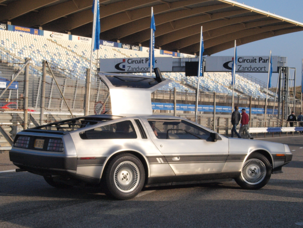 Ein silberner DeLorean DMC-12 fährt auf der Straße, umgeben von Menschen, Fahnen, Schildern, Geländern, Stühlen und einer Hütte, mit Bäumen und einem klaren blauen Himmel im Hintergrund.