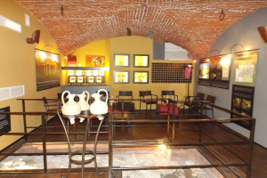 Ein Museumsraum voller Tische, Stühle, einem Geländer, Vasen, Texttafeln, Wandrahmen, Lampen und diversen Ausstellungsstücken.