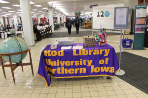 Eine Ausstellung mit einem Tisch, auf dem verschiedene Landesflaggen, eine Box, Papiere und ein Banner sowie links daneben eine Weltkugel, rechts daneben eine Anschlagtafel mit einigen Menschen in der Nähe und mehrere Tische und Stühle im Hintergrund zu sehen sind.