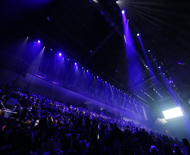 Eine große Menschenmenge, die in einem Konzertsaal auf einer Bühne sitzt, mit Deckenleuchten und einem Bildschirm auf der rechten Seite.