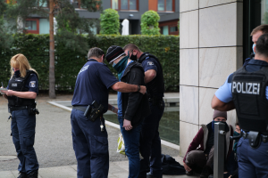 Eine Gruppe von Polizeibeamten steht vor einem Gebäude, eine Frau hält ein Buch und einen Stift in der Hand, während einige Menschen stehen und sitzen und Pflanzen, Bäume und Gebäude im Hintergrund zu sehen sind.