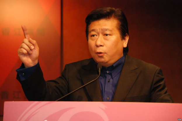 Ein Mann in Anzug und Krawatte, der vor einem Rednerpult mit Mikrofon spricht und ernst wirkt.