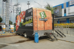 Ein weißer Lastwagen vor einem Gebäude mit einer Tafel, auf der "Teilen Sie eine positiv überladene Erfahrung" steht, mit Stufen, einem Zylinder am Boden, Geländern, Strommasten mit Drähten, Laternenmasten, Bäumen und einem bewölkten Himmel im Hintergrund.