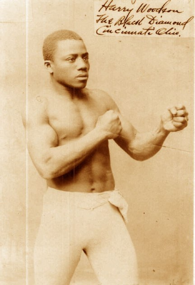 Altes Schwarz-Weiß-Foto eines Boxers im Ring mit Handschuhen und Shorts, der mit konzentrierter Miene und entschlossener Haltung steht.