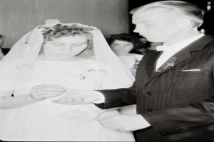 Ein Schwarz-Weiß-Foto einer Braut in einem weißen Kleid und Schleier und eines Bräutigams in einem Anzug beim Austausch von Ringen, mit einigen Menschen im Hintergrund.