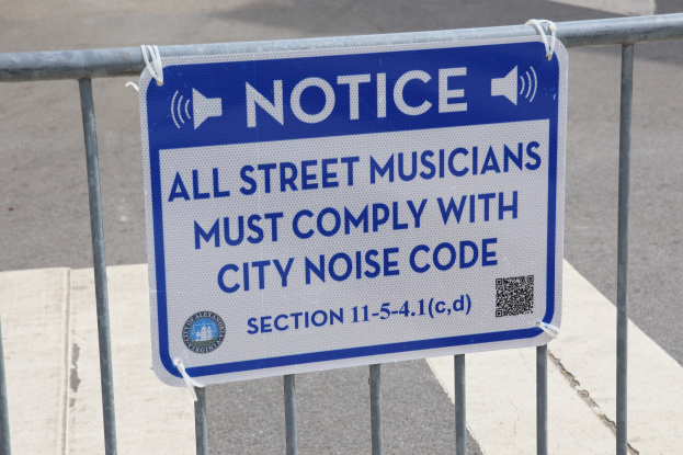 Anschlagtafel an einem Zaun mit einem Schild, auf dem "Anschlag: Alle Straßenmusiker müssen die Stadtlärmvorschriften einhalten" steht und eine Straße im Hintergrund.