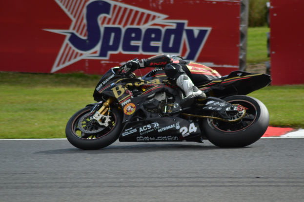 Eine Person mit Helm und Schuhwerk fährt ein Motorrad auf einer Rennstrecke, mit Gras und einer Tafel mit Text im Hintergrund.