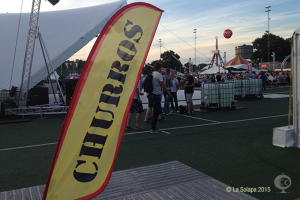 Ein lebhaftes Karnevalszelt mit Zelten, einem zentralen Mast, Menschen, Bäumen, Strommasten, Straßenbahnen auf der Straße und einem Eisenständer auf der linken Seite unter einem klaren Himmel.