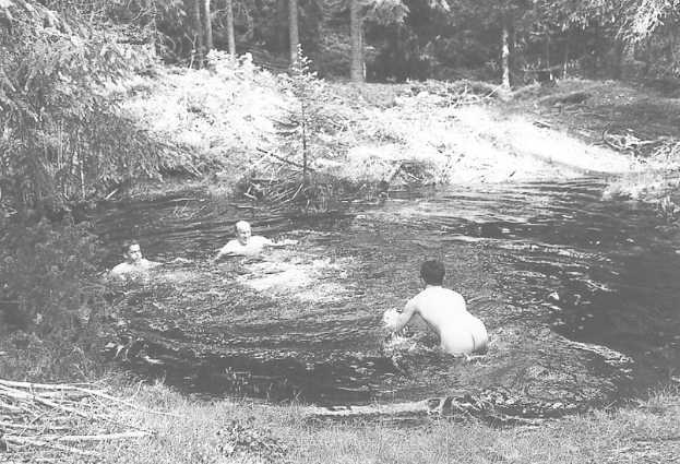 Schwarzes und weißes Foto von Menschen, die in einem Fluss umgeben von Bäumen und Gras schwimmen.