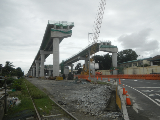 Baustelle mit einer Brücke im Hintergrund, Straße mit Verkehrskegeln markiert, Eisenbahnschiene links, verstreute Steine und Gras, Bäume und Gebäude auf beiden Seiten und ein bewölkter Himmel.