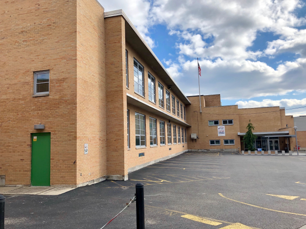 Ein großes Backsteingebäude mit einer grünen Tür und Fenstern, ein Flaggenmast mit einer wehenden Flagge davor, Bäume, Pfosten und ein bewölkter Himmel, das neue Schulgebäude an der University of North Dakota darstellend.