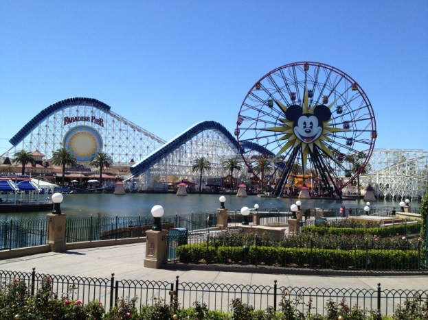 Der Disneyland California Adventure Freizeitpark mit einem Riesenrad und einer Achterbahn, umgeben von Grünflächen und Laternenmasten, mit Wasser im Vordergrund unter einem blauen Himmel.