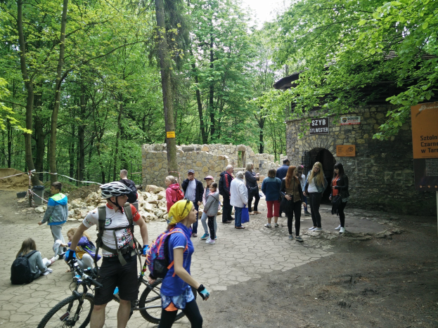 Gruppe von Menschen, die einen Waldweg entlanggehen, einige tragen Helme und tragen Taschen oder Fahrräder, mit einer Steinmauer und einem Schild im Hintergrund, das den Eingang zum Szyb Stylweste Mountainbike Trail anzeigt.