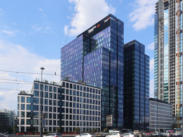 Außenansicht des Deutschen Bank-Hauptsitzes in Frankfurt, Deutschland, mit einem großen mehrfenstrigen Gebäude, umgeben von städtischer Infrastruktur wie Straßenlaternen, Fahrzeugen, Grünflächen und einem bewölkten Himmel.