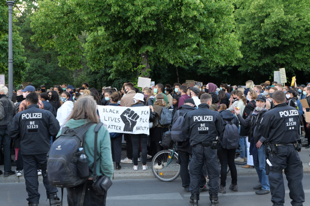 Eine große Gruppe von Menschen nimmt an einer Black Lives Matter Demonstration in Berlin teil, einige halten Schilder und andere tragen Kappen und Taschen, während im Vordergrund ein Fahrrad und im Hintergrund Bäume und ein Pfahl zu sehen sind.