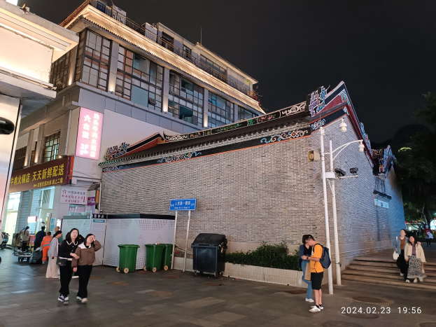 Menschen auf einer nächtlichen Straße mit Gebäuden, Straßenlaternen, Schildern, Pflanzen und Bäumen, einige mit Taschen.