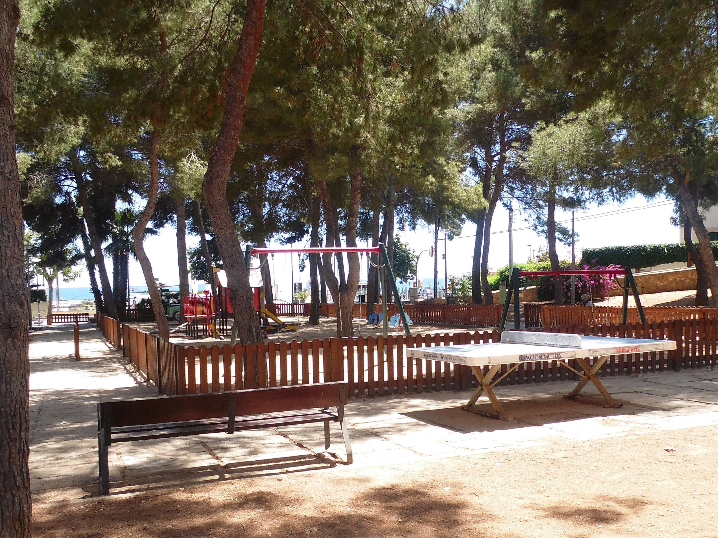 Parkszene Mit Zentralem Tischtennis-Tisch, Bänken, Schaukeln, Pfählen, Pflanzen Und Bäumen, Mit Einem Zaun Und Einem Wasserbecken Im Hintergrund Unter Einem Klaren Blauen Himmel.