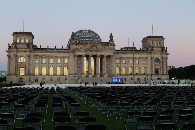 Das Reichstagsgebäude in Berlin, Deutschland, mit seiner markanten Kuppel, umgeben von Menschen, Stühlen, Straßenlaternen, Schildern, Bäumen und einem bewölkten Himmel.