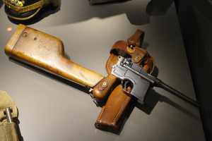 Eine Luger-Halbautomatik-Pistole in einer Glasvitrine ausgestellt, begleitet von einer Mütze und anderen Gegenständen auf einem Tisch.