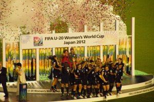 Gruppe von Frauenfußballspielerinnen in schwarzen Uniformen, die auf einer Bühne feiern und von einer Menge mit Kameras umgeben sind, mit einem Hintergrund aus Text, Bildern und Dekoration auf Gras.