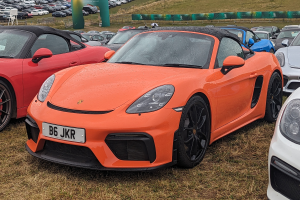Ein Porsche 718 Boxster GTS parkt auf einer grünen Fläche beim Goodwood Festival of Speed, zusammen mit anderen Autos, einer Fahne mit Text, Bäumen und einem klaren blauen Himmel im Hintergrund.