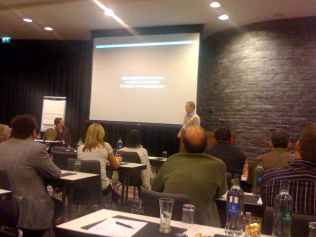 Eine Gruppe von Menschen hört einem Mann zu, der mit einem Bildschirm hinter sich präsentiert.