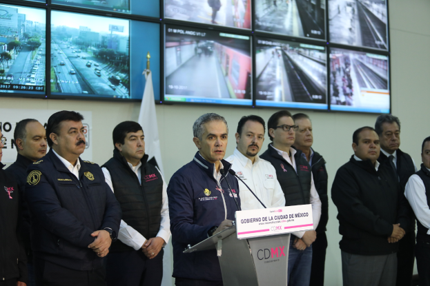 Eine Gruppe von Männern vor einem Podium mit einem Mikrofon und einer Tafel mit der Aufschrift "Mexikanische Zoll- und Grenzschutzbehörde", mit Fernsehgeräten und einer Flagge im Hintergrund.