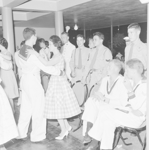 Schwarz-weiß-Bild von Militärpersonal, das tanzt und sich vergnügt in einem dekorierten Saal mit Säulen und Beleuchtung.