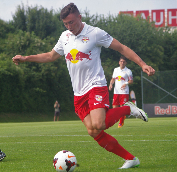 Ein professioneller Fußballspieler in einem roten Trikot kickt einen Ball auf einem grünen Feld mit Bäumen und einem klaren blauen Himmel im Hintergrund, mit "RB Leipzig" auf einer Tafel.