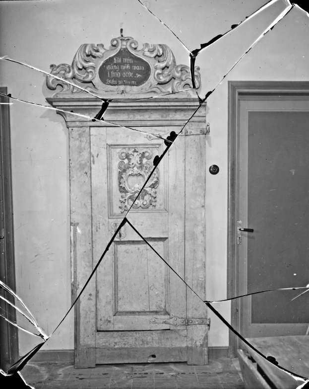 Ein Schwarz-Weiß-Foto einer zerbrochenen Tür in der Mitte eines Raumes, mit zerbrochenem Glas drumherum und einer Wand im Hintergrund.