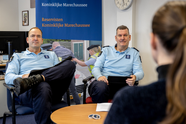 Zwei Polizeibeamte sitzen an einem Tisch mit einer Frau, Papieren und Gegenständen auf dem Tisch, einer Wandbanner und Uhr im Hintergrund, einem weiteren Tisch mit Monitor, Tastatur, Maus und Gegenständen links daneben und Fotorahmen an der Wand.