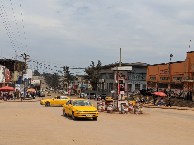 Eine belebte Stadtstraße mit einem gelben Taxi, das fährt, Passanten mit Regenschirmen auf den Gehwegen, Gebäude mit Fenstern und Strommasten mit Drähten unter einem bewölkten Himmel.