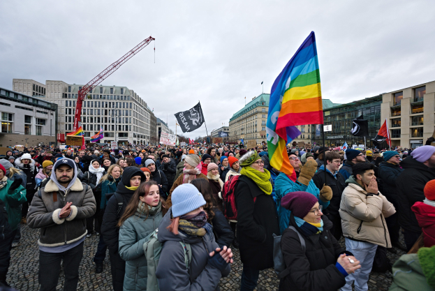 Eine große Gruppe von Menschen steht vor einem Gebäude und hält Fahnen und Schilder mit der Aufschrift "Lgbtq+ Rechte Demonstration in Berlin", einige tragen Mützen und tragen Taschen, während Gebäude, ein Kran und ein bewölkter Himmel im Hintergrund zu sehen sind.