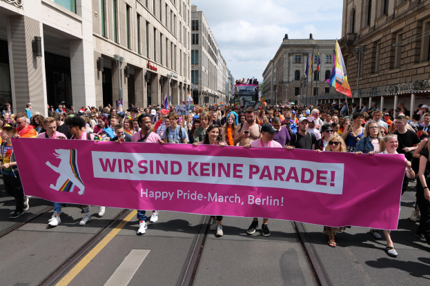 Eine Gruppe von Menschen marschiert auf einer Straße in Berlin und hält ein pinkes Banner mit der Aufschrift "Happy Pride March".