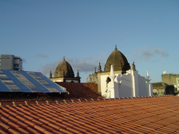 Stadtansicht mit Gebäuden im Vordergrund, Solarpanels auf einem Dach und einem klaren blauen Himmel im Hintergrund.