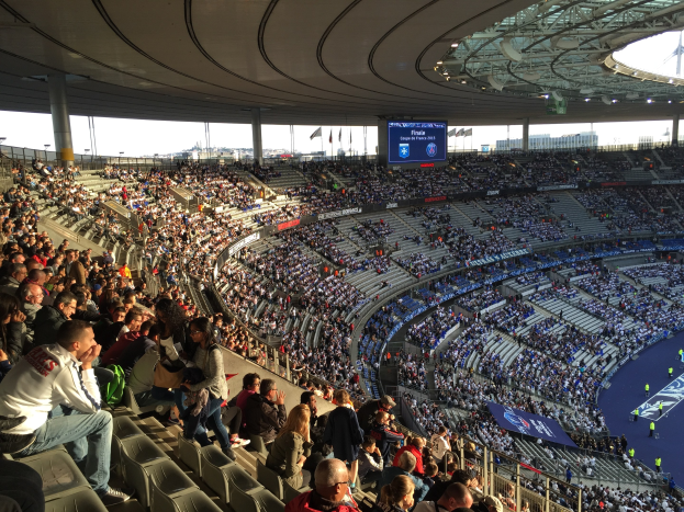 Große Menge in einem Stadion bei einem Fussballspiel, mit einer Bühne rechts, Fahnen, Stangen, einem Bildschirm und dem Allianz Stadion in München im Hintergrund.