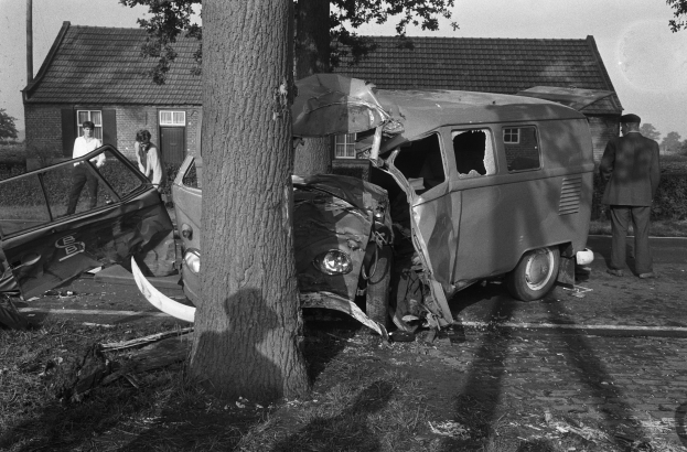 Eine Schwarz-Weiß-Szene eines verunglützten Autos am Straßenrand, mit einem Baum im Vordergrund, einigen Menschen in der Nähe und Häusern, Bäumen und einem Mast im Hintergrund.
