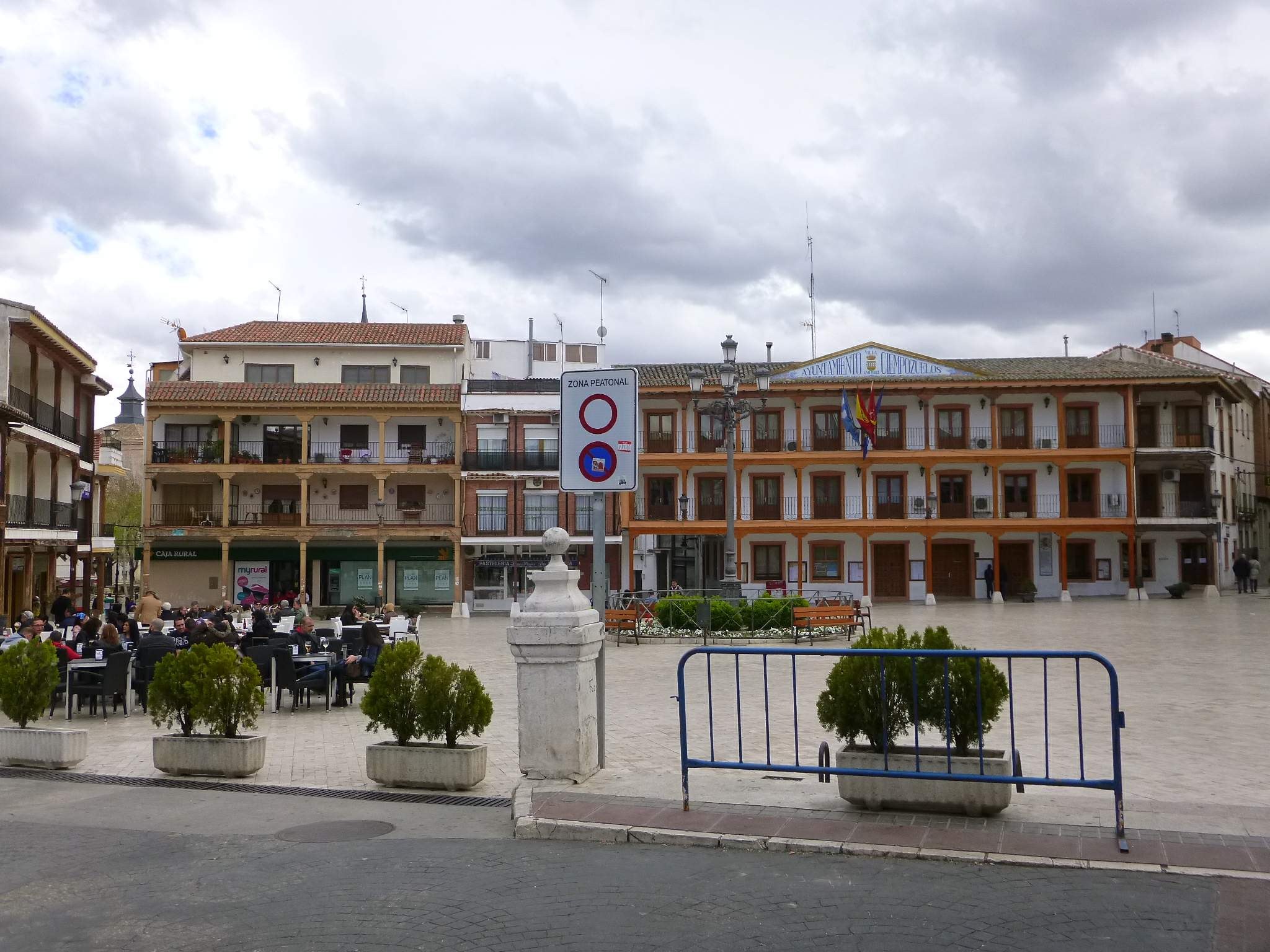 Ein belebter Stadtplatz mit Menschen, die sitzen und stehen, Topfpflanzen, Metallabsperrungen, ein Schild an einem Pfahl, Straßenlaternen mit Flaggen, umliegende Gebäude und ein bewölkter Himmel.